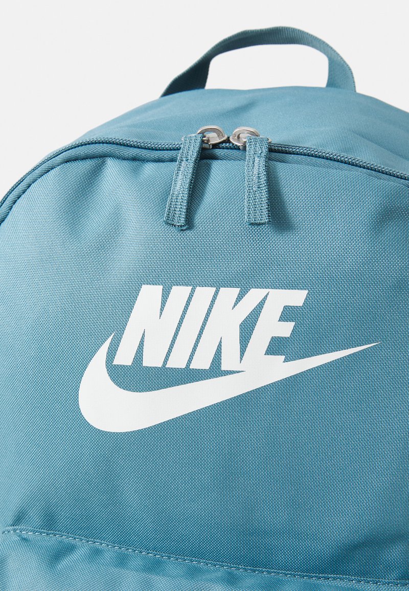 Mochila de nailon en color verde azulado con doble cremallera, que presenta un destacado logo de Nike en blanco y un asa superior resistente. Textura suave y diseño ligero.