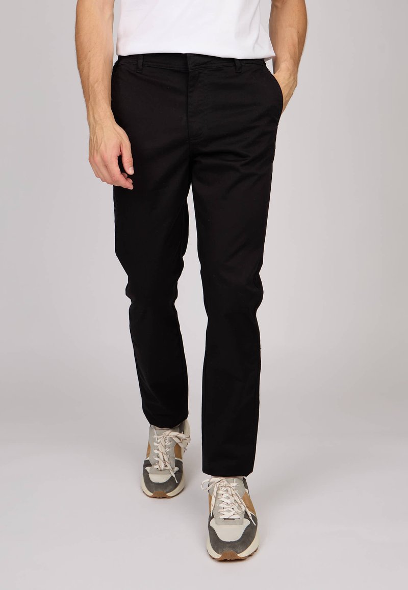Chino extensible noir à coupe slim, doté de poches latérales et d'une finition lisse, porté avec des baskets multicolores.