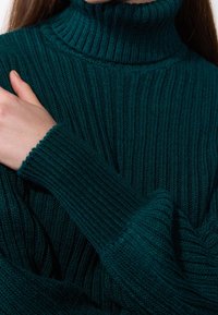 Kvinna som bär en mörkgrön ribbad polotröja, håller sin vänstra arm med sin högra hand, närbild på överkropp och händer.