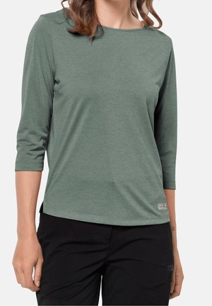 Vrouw die een groen shirt met lange mouwen en een zwarte broek draagt, staand met ontspannen armen langs haar zij tegen een effen achtergrond.
