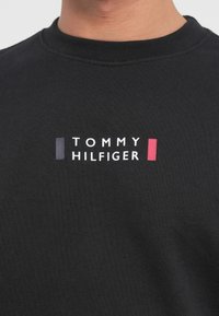 Μαύρο βαμβακερό φούτερ με λευκή λογότυπο "TOMMY HILFIGER" και κόκκινες και μπλε ορθογώνιες τονιστικές γραμμές κάτω από τα γράμματα.