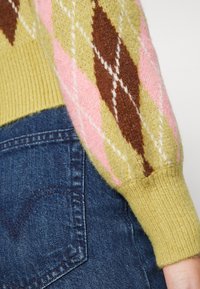 Stickad tröja med ett argyle-mönster i gult, rosa och brunt; ribbade ärmslut; tillsammans med mörk denimjeans som har detaljer på fickorna.