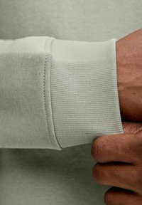 Jasnozielony mankiet długiego rękawa, o prążkowanej fakturze, z gładkim wykończeniem i starannym szwem, trzymany przez brązową rękę.