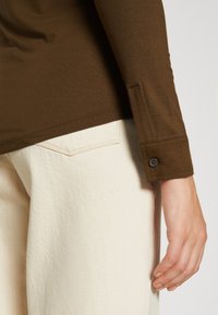 Top marrón de manga larga con diseño ajustado y puños con botones, combinado con pantalones blancos texturizados que presentan detalles de costuras en contraste.