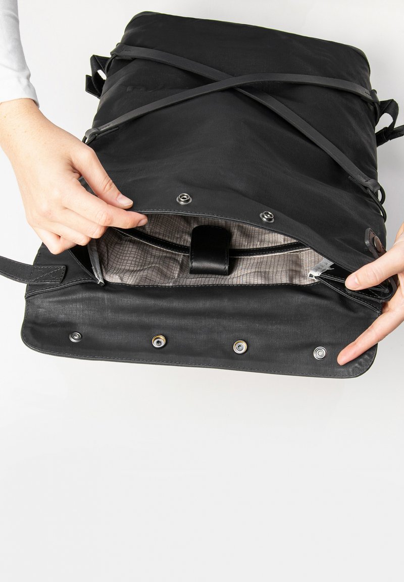 Zwarte tas met een stijlvolle textuur, voorzien van een beveiligde opening, zilverkleurige hardware en een gemêleerd interieur dat zichtbaar is vanuit handen die de klep vasthouden.