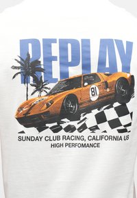 Oranžs sacīkšu auto ar numuru 81 virs melnbaltā šaha karoga, palmu kokiem un lieliem ziliem tekstiem "REPLAY".