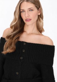 Pulover negru din tricot cu umerii descoperiți, având o textură cu nervuri, cu butoane mari de culoare închisă pe față și manșete largi.