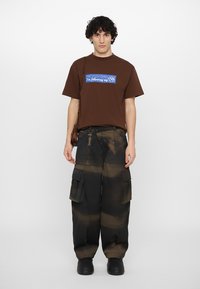 Brown cotton t-shirt with blue text graphic, paired with black cargo pants featuring a faded brown pattern and side pockets. Black footwear completes the look.