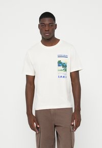 Kortermet kremfarge t-skjorte med grafisk trykk av et landskap på framsiden. Stoffet er bomull med en glatt tekstur.