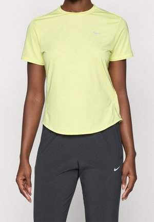 Osoba nosící světle žluté sportovní tričko s krátkým rukávem a tmavě šedé kalhoty, obojí s malými bílými logy Nike.