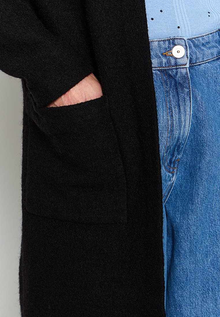 Schwarze Strickjacke aus weichem Material mit einer seitlichen Tasche, kombiniert mit einem hellblauen Oberteil und blauen Jeans, die einen Knopf am Bund haben.