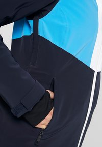 Bunda v blokovém vzoru námořnické modré a nebeské modré barvy má kapsu na zip, elegantní design a měkkou látku s matným povrchem.