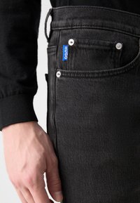Black denim jeans with a slim fit, featuring a visible blue "HUGO" label and silver hardware on the front pocket. Textured fabric close-up.
