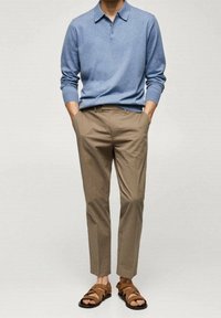Persona che indossa una polo a maniche lunghe blu, pantaloni sartoriali color marrone chiaro e sandali in pelle marrone, in piedi con le mani nelle tasche.