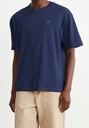 Mand iført en ensfarvet mørkeblå T-shirt med korte ærmer med et lille Timberland-logo på brystet og beige bukser.
