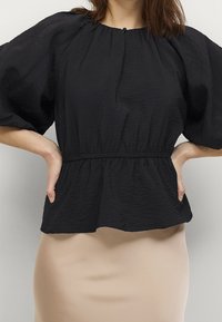 Black textured blouse featuring wide puff sleeves and an elastic waist, complementing a fitted, beige skirt. Casual yet refined design.