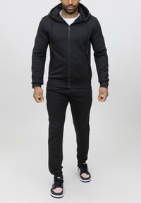 Haut à zip noir avec une finition texturée, associé à un pantalon de jogging noir assorti. Porté avec des baskets noires et roses avec une semelle blanche.