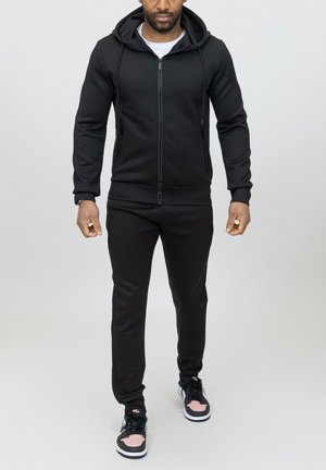 Haut à zip noir avec une finition texturée, associé à un pantalon de jogging noir assorti. Porté avec des baskets noires et roses avec une semelle blanche.