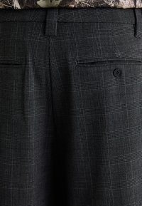 Pantalons à carreaux gris foncé avec une poche arrière boutonnée et des passants de ceinture, portés avec un haut camouflage.
