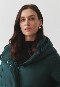 Zelená puffová bunda se zipem a velkou kapucí, vyrobená z prošívaného, lesklého materiálu. Model má pod bundou černý svetr.