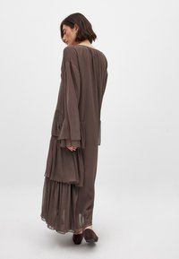 Lange bruine jurk met gelaagde ruches, doorschijnende textuur en brede mouwen. Zachte stof valt elegant tot de vloer, gecombineerd met donkere schoenen.