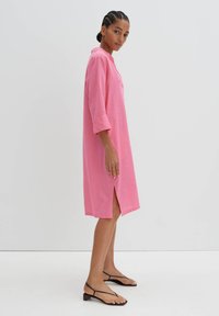 Rosa Leinenkleid mit 3/4-Ärmeln, Kragen, seitlichen Schlitzen und lockerer Passform. Kombiniert mit schwarzen Sandalen mit Riemchen. Schlicher weißer Hintergrund.