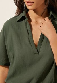 Blusa verde oliva de manga corta con escote en V. Tejido suave y texturizado con costuras visibles a lo largo del cuello y el dobladillo.