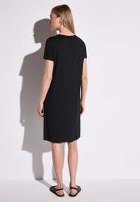 Schwarzes kurzärmeliges Kleid mit geradem Schnitt, aus weichem Stoff, mit rundem Ausschnitt und bis zur Mitte des Oberschenkels reichend. Getragen mit schwarzen Sandalen.