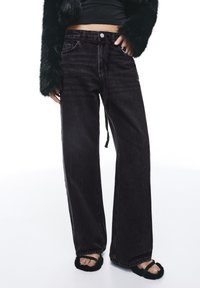 Jean en denim noir à jambes larges, taille haute, doté d'une braguette à boutons, d'une légère décoloration et d'une coupe ample. Associé à des sandales sombres et pelucheuses.