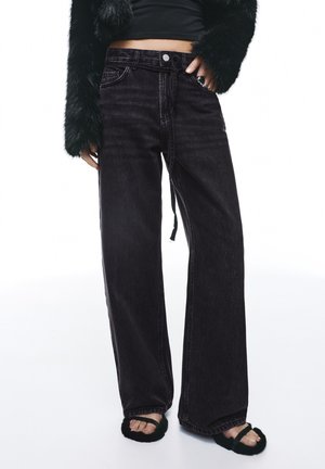 Črne široke kavbojke iz jeansa z visokim pasom, s patentnim zapiranjem na gumbe, z nežnim bledenjem in ohlapnim krojem. Kombinirane z mehko temno odprto obuvalo.