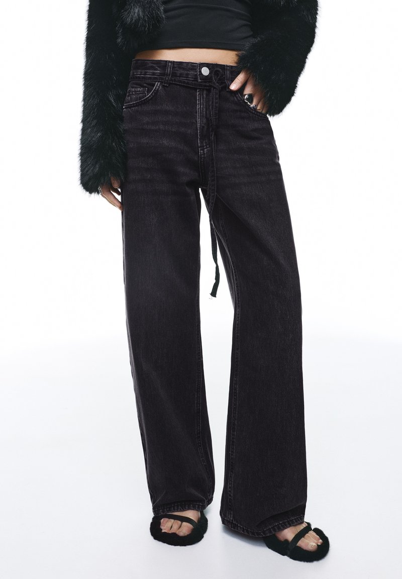 Črne široke kavbojke iz jeansa z visokim pasom, s patentnim zapiranjem na gumbe, z nežnim bledenjem in ohlapnim krojem. Kombinirane z mehko temno odprto obuvalo.