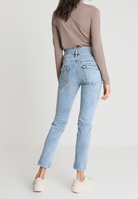 Světle modré džíny s vysokým pasem a úzkým střihem, s pěti kapsami a vybledlou texturou. V kombinaci s hnědým pruhovaným topem s dlouhým rukávem a světlými teniskami.