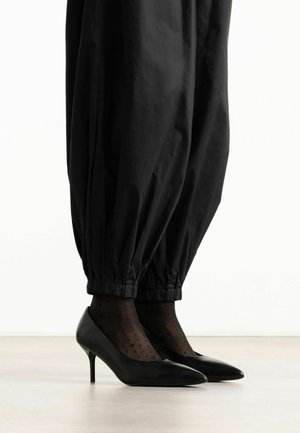 Benen met zwarte taps toelopende broeken met elastische boorden, doorschijnende zwarte sokken met stippen en puntige zwarte hoge hakken op een lichte vloer.