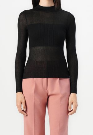 Personne portant un pull noir côtelé à manches longues et un pantalon rose taille haute sur mesure, debout sur fond blanc.