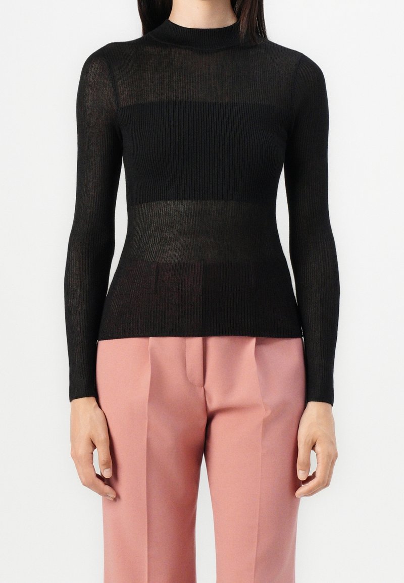 Personne portant un pull noir côtelé à manches longues et un pantalon rose taille haute sur mesure, debout sur fond blanc.