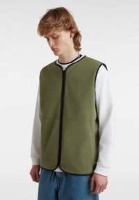 Gilet in pile verde con zip e bordi neri, dotato di due tasche laterali e un orlo arrotondato, indossato sopra una camicia a manica lunga bianca.
