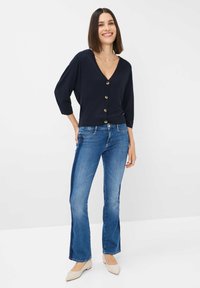Camicetta blu navy con scollo a V, abbinata a jeans chiari a campana e scarpe beige a punta, su sfondo bianco.
