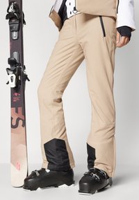 Beige snø bukser med rillet tekstur, svarte detaljer ved mansjettene, kombinert med svarte skistøvler. En ski er plassert ved siden av.
