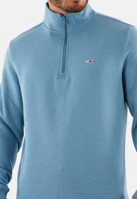 Pullover a mezza zip di colore azzurro chiaro realizzato in tessuto morbido. Caratterizzato da un colletto alto e un piccolo logo sul petto. Texture liscia con maniche lunghe.
