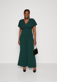 Groene jumpsuit met een V-hals, korte wijdvallende mouwen, passige taille en wijde broekspijpen. Gekwens met een kleine zwarte handtas en puntige hakken.