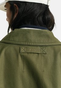 Olivegrüner Jacke aus strukturiertem Material, mit einem navyblauen gerippten Kragen und GANT-Logopatch auf dem Rücken. Sichtbare Steppdetails.