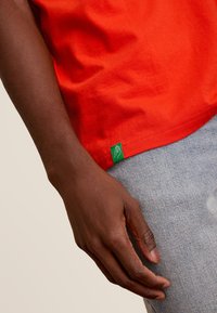 Rode katoenen t-shirt met korte mouwen en een groene label aan de zoom. Een hand houdt het shirt vast, tegen een gestructureerde lichtblauwe denim achtergrond.