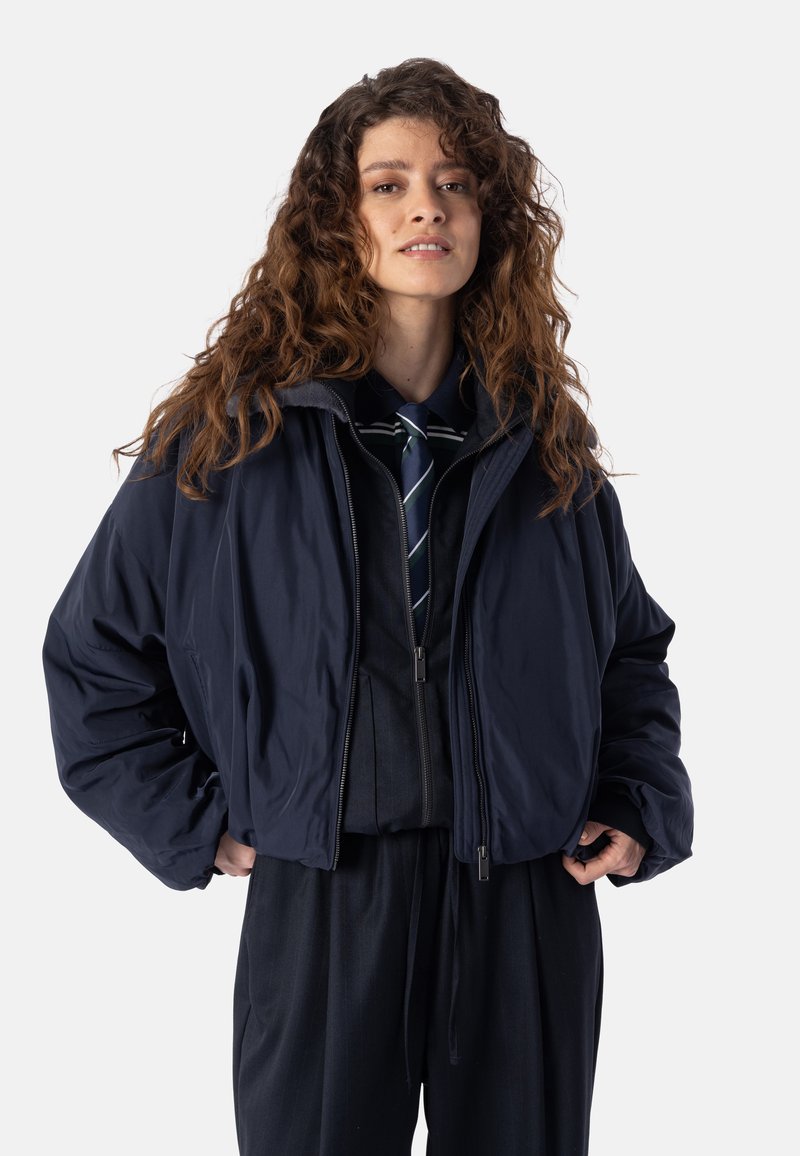 Jeune femme aux cheveux bouclés portant une veste bleu marine foncé par-dessus un pull zippé et une cravate rayée, debout les mains dans les poches de la veste.