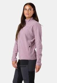 Vaaleanpunainen fleece-takki, jossa on korkea kaulus, etuvetoketju ja sivutaskut. Takissa on pehmeä pinta ja brodeerattu logo rinnassa.