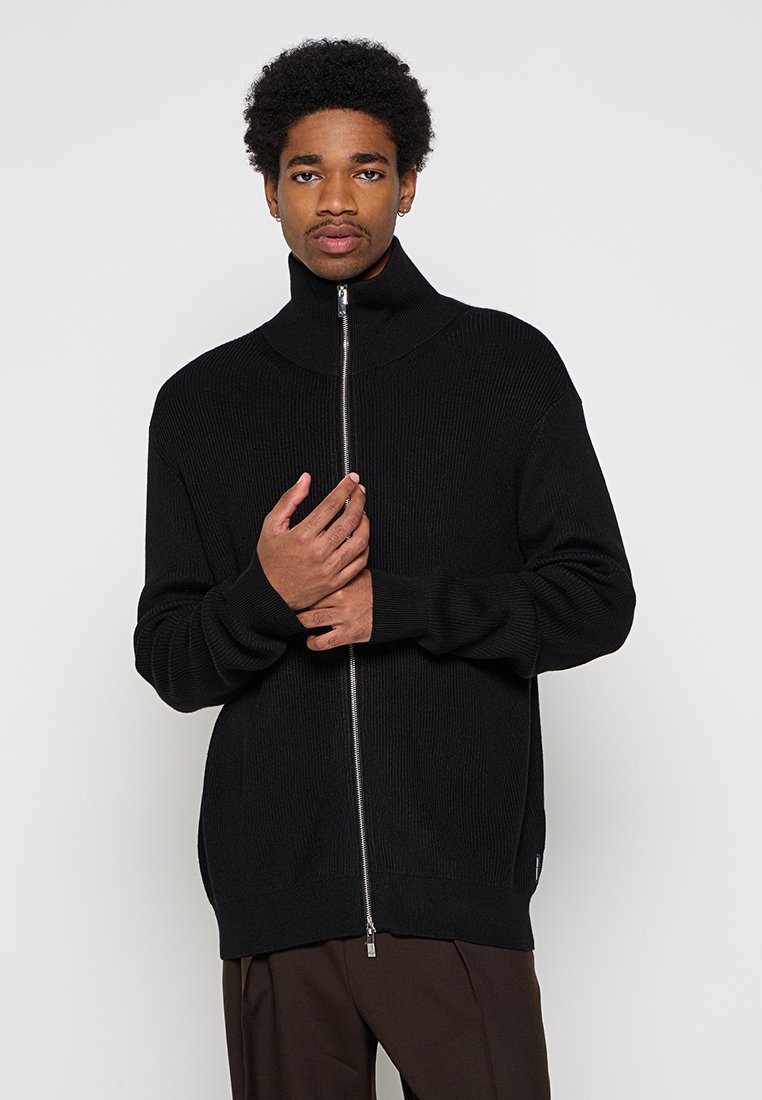 Schwarzer gerippter Reißverschluss-Pullover mit hohem Kragen, langen Ärmeln und einem Frontreißverschluss. Der Stoff hat eine glatte Textur und einen lockeren Schnitt.