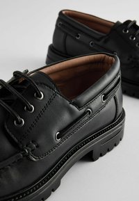 Zapatos de cuero negro con una textura suave, punta redonda y una suela gruesa y robusta. Presentan un diseño de cordones y ojales de plata.