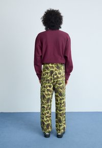 Bordeaux sweatshirt en losvallende groene camouflagebroek, met meerdere zakken, gestructureerde stof en zwarte schoeisel. Achtergrond is lichtblauw.