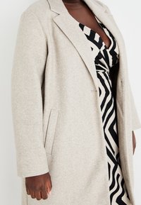 Beżowy płaszcz z mieszanki wełny z klapami z wycięciem, długimi rękawami i bocznymi kieszeniami, noszony na czarno-białej sukience w wzory.