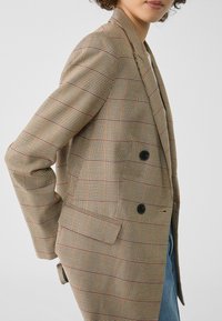 Bruine geruite dubbelbreasted blazer met contrasterende rode lijnen, geknikte revers en twee zwarte knopen. Stof lijkt glad met een gestructureerde pasvorm.