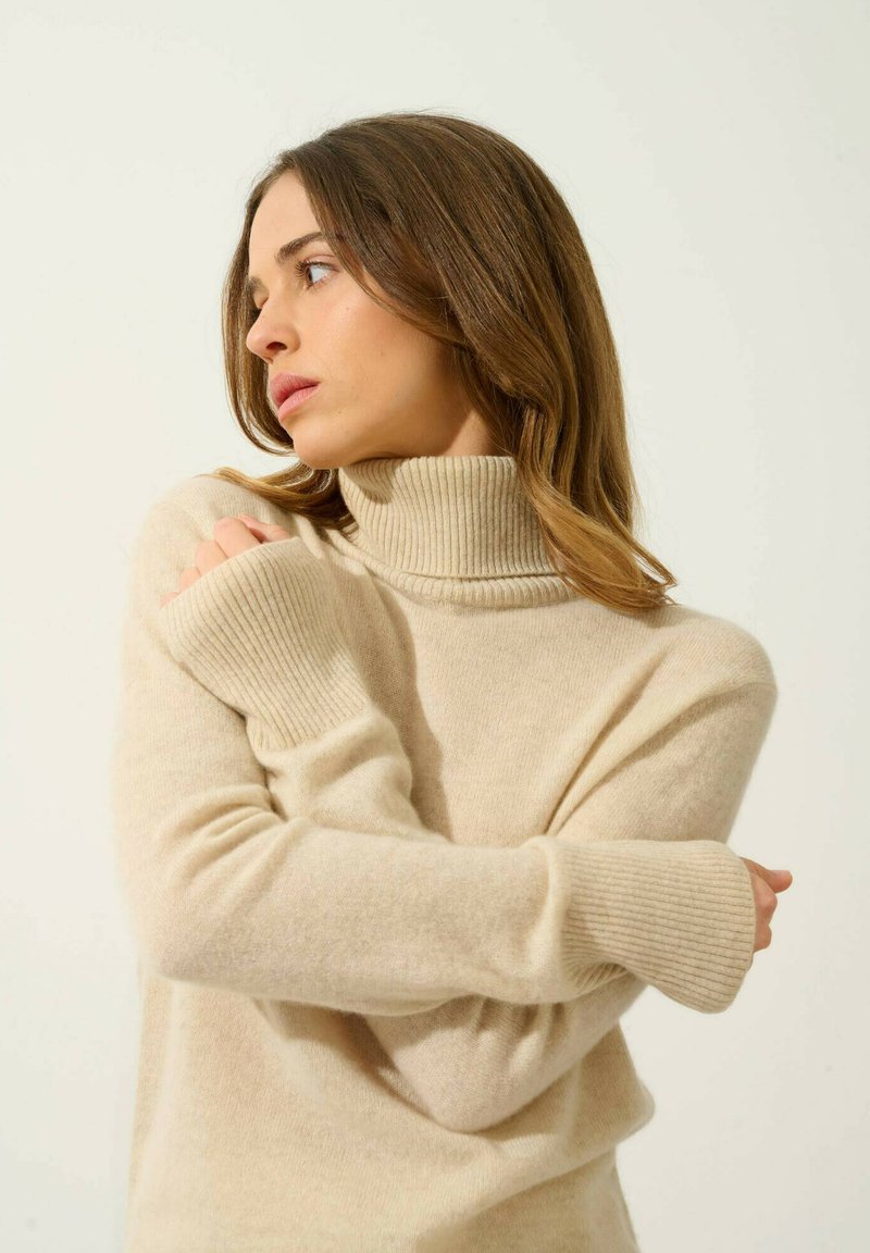 Beżowy sweter z golfem, z prążkowanymi mankietami i kołnierzem, wykonany z miękkiej, teksturowanej tkaniny, o luźnym kroju i długich rękawach.